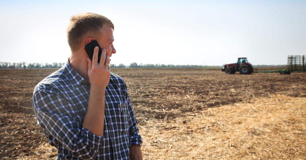 Young man with phone in a field and a tractor on a background.