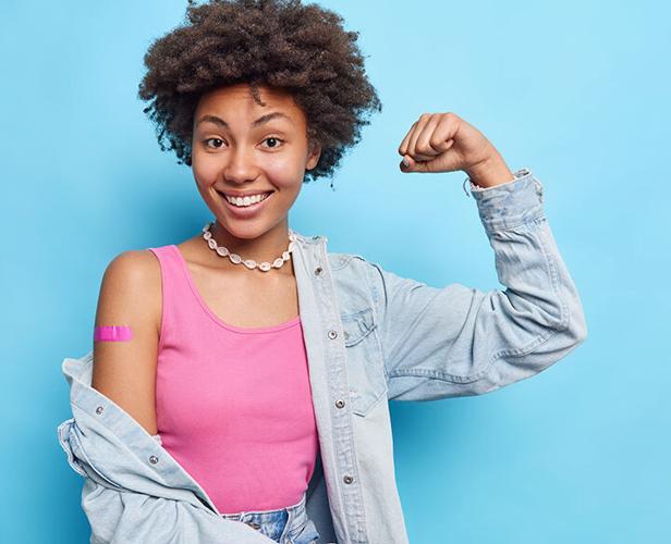 Photo of satisfied curly haired woman raises arm shows biceps wears pink t shirt denim shirt necklace adhesive bandage on shoulder after vaccination feels good and protected encourages to vaccinate
