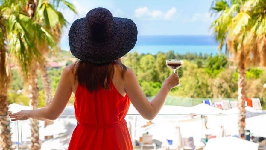 Back view of Woman in a red dress and beach hat holding cold coffee drink frappe enjoys the summer sea view, overlooking the Mediterranean Sea in Italy, Sicily. No visible face. Vacation.