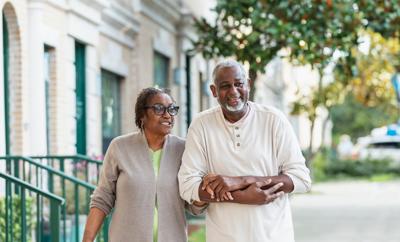 A senior African-American couple holding hands as they walk outdoors, by a building on a city sidewalk. They are in their 70s, smiling and relaxed.