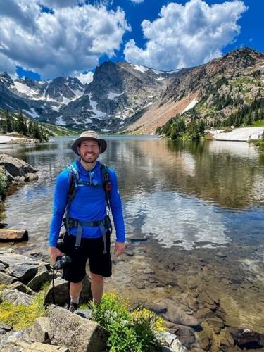 Hiker in blue jacket standing at the edge of a clear alpine lake with snowy peaks