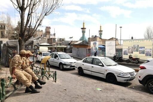 Two Iranian soldiers look at their mobile phone as they sit close to an image of Iran's slain supreme leader Ayatollah Ali Khamenei (R), along a street close to Tajrish Square in Tehran on March 7, 2026