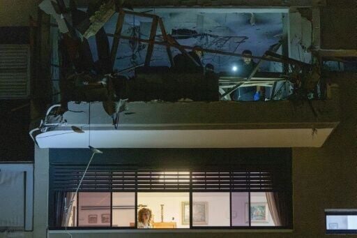 A woman stands at a window as first responders and security forces inspect a damaged apartment above after the building was hit by an Iranian missile strike in Ramat Gan, on the outskirts of Tel Aviv, early on March 18, 2026
