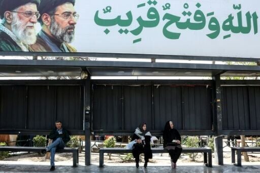 Iranians wait at a bus stop as they sit under a banner featuring Iran's late supreme leaders Ayatollah Ali Khamenei (L) and his son newly elected supreme leader Mojtaba Khamenei, in northern Tehran