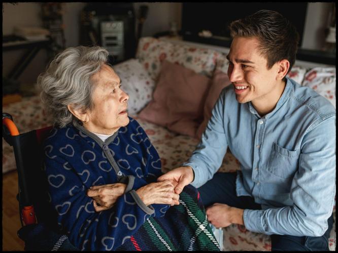 Young man sitting next to an old sick aged woman in wheelchair taking her hands while talking and smiling