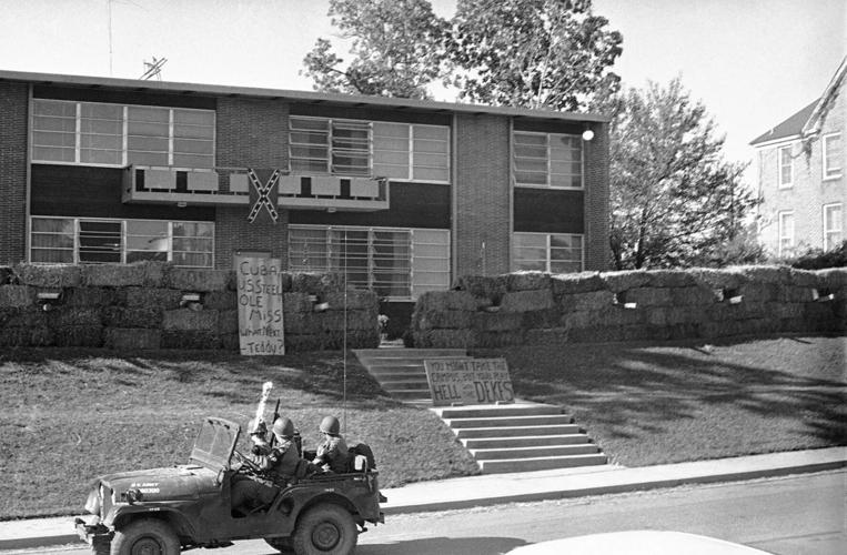 A black and white photo of a military jeep driving past a campus fraternity house surrounded by a hay bale wall