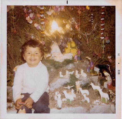 A young boy with medium-tone skin sits next to a Nativity scene. He's wearing a white shirt and dark trousers.