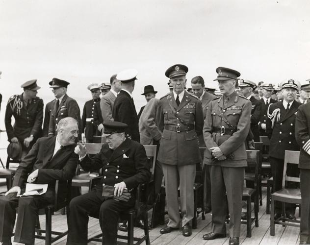 This black and white photograph is of President Franklin D. Roosevelt (seated left) and British Prime Minister Winston Churchill (seated right) in a candid moment aboard the HMS Prince of Wales during the Atlantic Conference in Placentia Bay, Newfoundland.