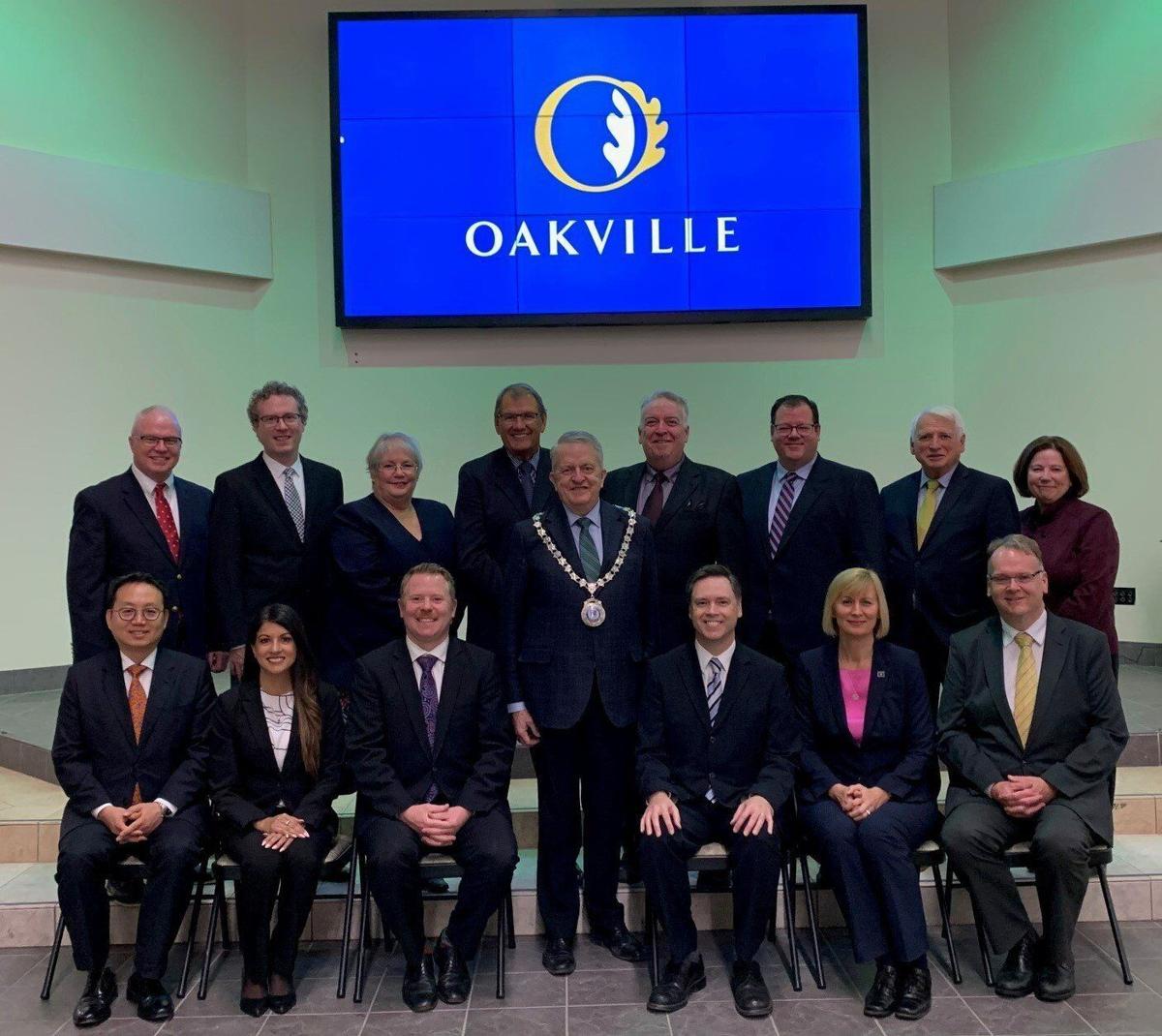 Oakville council, front row sitting, back row standing, with Mayor Rob Burton standing in the middle. Behind them, an Oakville logo is projected onto a screen
