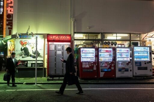 Two businessmen walk past advertisements and a row of vending machines in Tokyo's Shinjuku on April 13, 2026