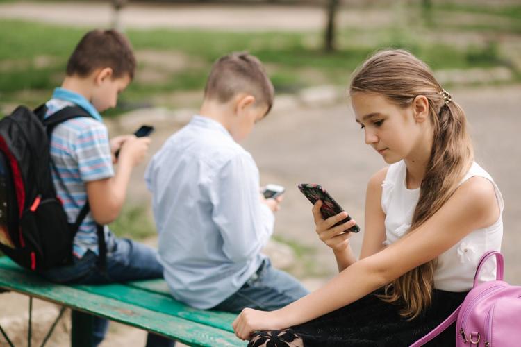 Two boys and girl use their phones during school breack. Cute boys sitting on the bench and play online games