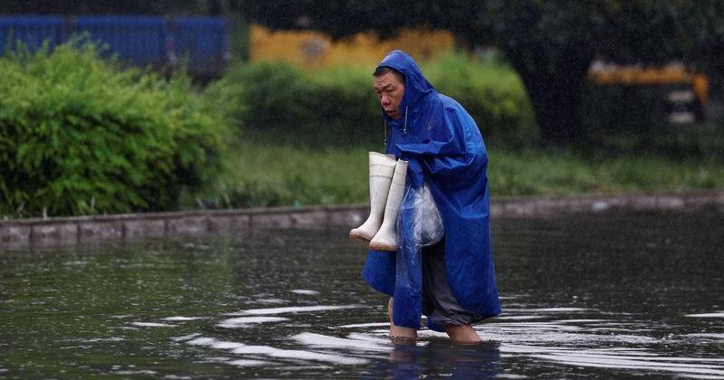 Man in raincoat holds a pair of rain boots as he wades through floodwaters on a street, amid heavy rainfall brought by remnants of Typhoon Dolphin, in Luohe