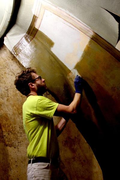 Emanuel Fritz of Conrad Schmitt Studios uses a paintbrush to apply cleaning solution to a 6” square in the painting. By gently applying and wiping away the cleaning agent, Fritz revealed the artistry beneath the grime.