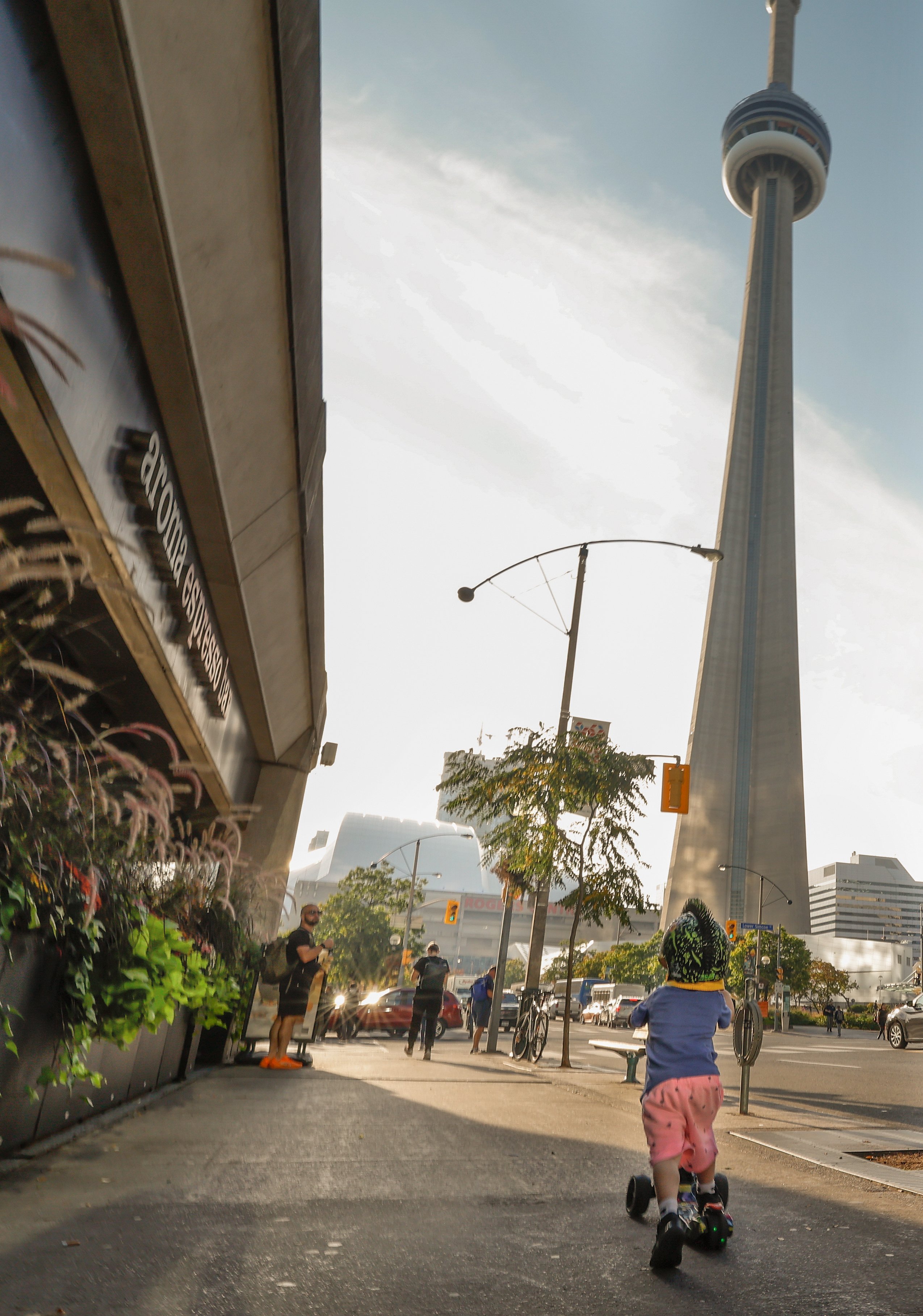 A young boy rides his scooter through a downtown sidewalk. The CN tower is visible in the background