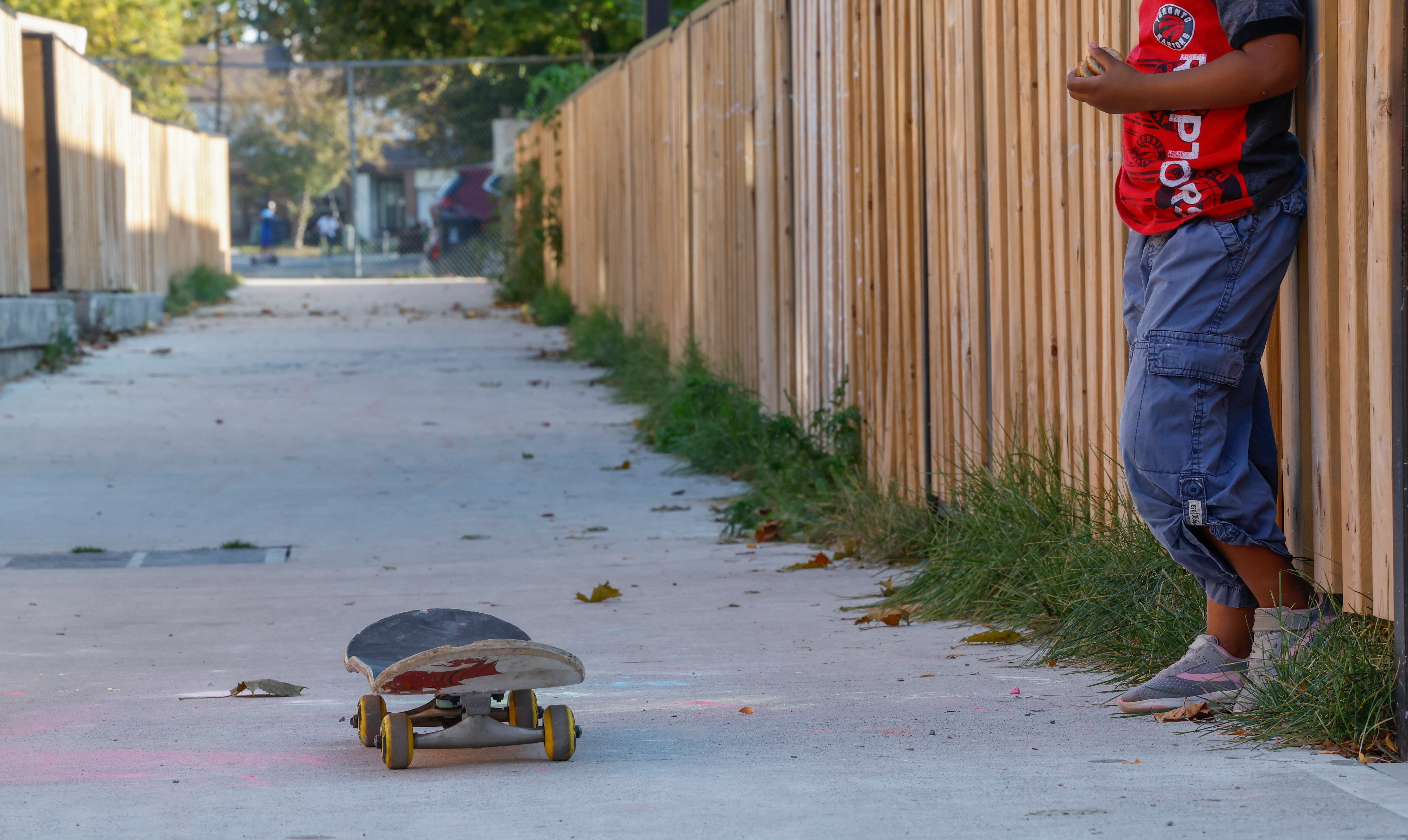 A photo of a young girl standing against a fence in a laneway. There is a skateboard on the ground in front of her