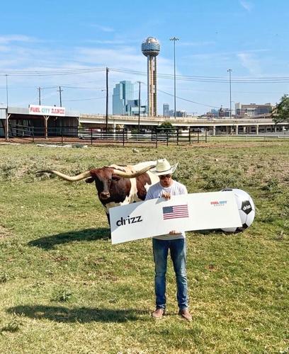 Drizz founder Rodrigo Ricaud, a Mexican-American entrepreneur from Dallas, holds a USA sign beside Buddy the Longhorn at Fuel City Ranch. Photo courtesy of Drizz (drizzdrops.com).