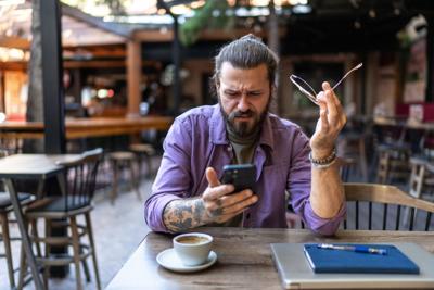 Young adult man feeling frustrated and confused while looking at his smartphone, holding his glasses, sitting at a table with coffee and laptop in an outdoor cafe