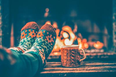 Feet in woollen socks by the Christmas fireplace. Woman relaxes by warm fire with a cup of hot drink and warming up her feet in woollen socks. Close up on feet. Winter and Christmas holidays concept.