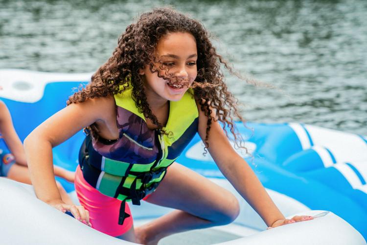 Smiling happy girl in life jacket with long curly hair playing on floaty on lake
