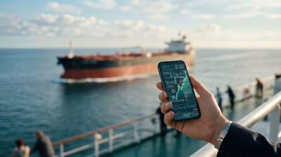 Hand holding smartphone displaying financial data with oil tanker ship navigating in the background on a clear day, showcasing global trade and energy logistics