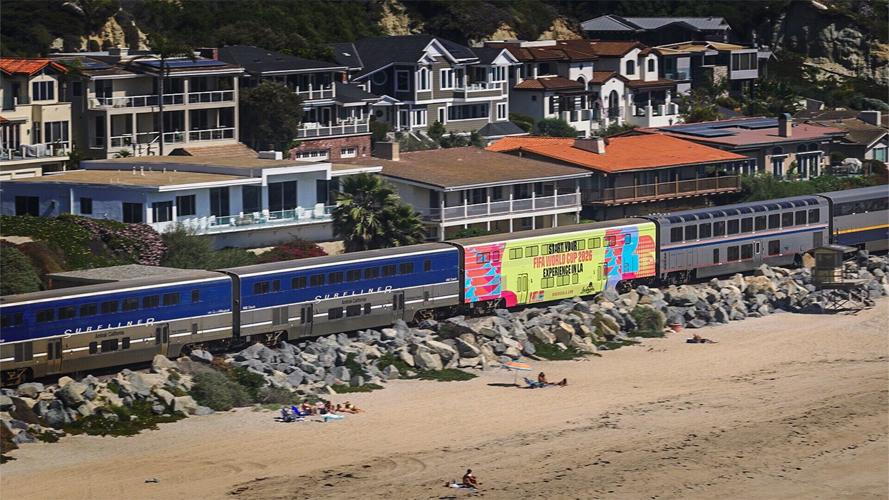 An Amtrak Pacific Surfliner train travels along the Southern California coastline featuring a special LA Tourism wrap promoting summer 2026 soccer matches in Los Angeles.