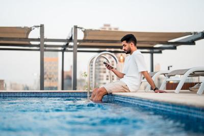Lifestyle portrait of young Middle Eastern man in white clothing sitting poolside at luxury rooftop terrace, browsing smartphone while enjoying peaceful urban views