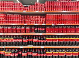 Photo of a fully stocked supermarket shelf filled with neatly arranged Coca-Cola cans and bottles in various sizes.