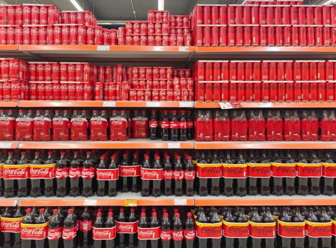 Photo of a fully stocked supermarket shelf filled with neatly arranged Coca-Cola cans and bottles in various sizes.